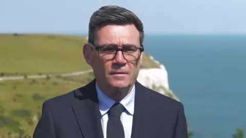 Mid shot of Andy Burnham wearing a black suit and black tie with black-framed glasses. In the distance is the white cliffs of Dover.