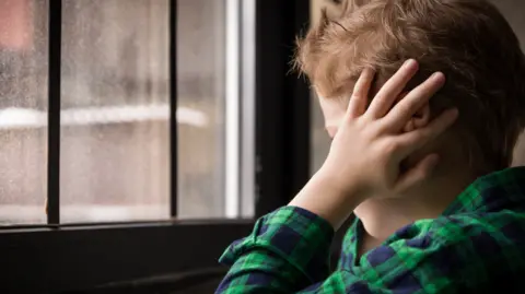 Getty Images Stock image of a child in a green and blue check shirt staring out of the window with their hands over their ears.  