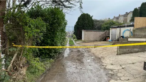 The crime scene in a lane off Dransfield Way, Bath, where the attack took place, it is a narrow muddy lane with garages on one side and bushes on the other