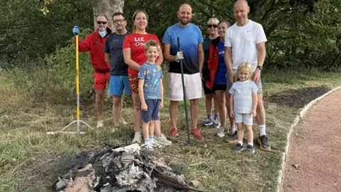 A group of nine people, three women, four men and two children, in shorts and t-shirts, some holding rakes, standing on the grass verge of a running track.