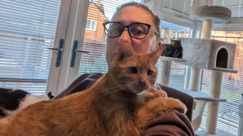 RSPCA Claire Craig is sitting in a chair with a short-haired ginger cat in her arms. There is a large cat climber behind her and another cat is sitting on that. A garden can be seen in the background behind blinds on the windows and patio doors