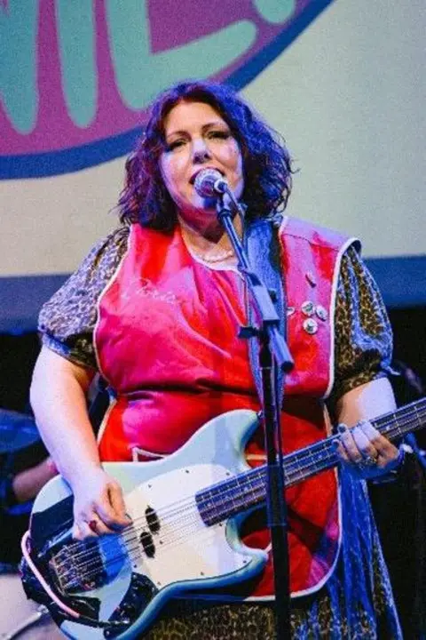 The NaNaz A woman is wearing holding a guitar on stage and is singing into a microphone.