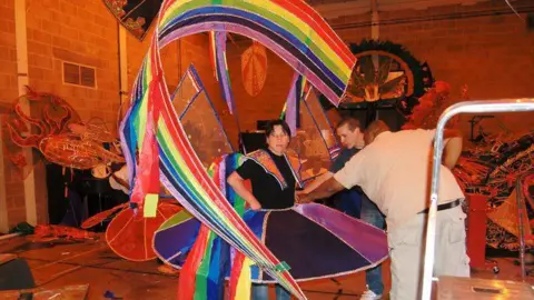 Matthew Catlin A man stands as two others adjust a large rainbow carnival costume on him