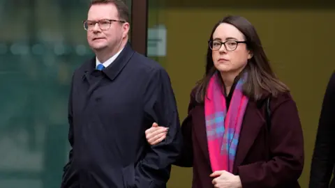 PA Media Conor McGinn, 41, who has short brown hair, glasses with tortoise-shell frames and is wearing a dark blue mackintosh coat, walks away from a court building alongside a woman with long brown hair, glasses, a red jacket and pink scarf. She is linking arms with McGinn and looks into the camera with a serious expression. 