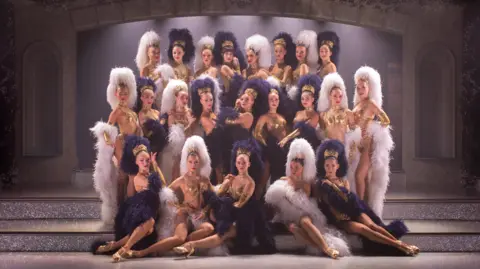 Thursford Christmas Spectacular A collection of 22 female dancers are organised in three rows, with the front row sitting and each row alternating between dancers in blue costumes and those in white, on a theatre stage with some scenery in the background.