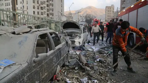View of damaged building, person cleaning up rubble next to a car