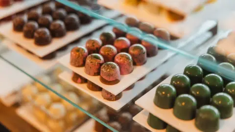 Jon Collins A selection of artisan chocolates on white serving plates in a display cabinet. The chocolates come in a range of colours, including green, pink and yellow.