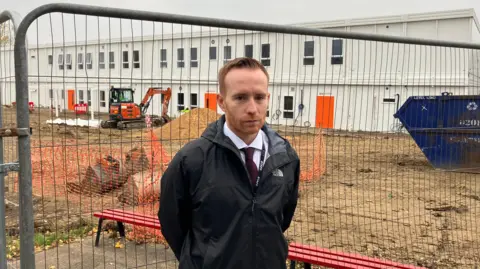 Matt Knight/BBC A head-and-shoulders picture of Katherines Head of School, Julien Mealey. He has short, red hair and a beard. He is wearing a black coat and a white shirt and black tie. He is standing in front of a fence. On the other side of the fence, there is a building site. A two-storey temporary building is being constructed.