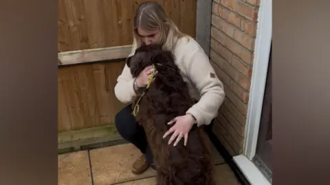 Contributed Rebecca Harvey crouches in the corner of a garden as she holds her dog flora. Rebecca has long blonde hair and wears a cream jumper with dark trousers and brown shoes. Flora has brown fur and a yellow lead on. 