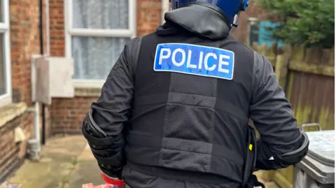 Cambridgeshire police A close-up of the back of a uniformed police officer. He is wearing a blue helmet and a black uniform and is walking down a path at the side of a house. On the back of his jacket a blue badge says "POLICE".