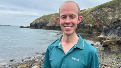 A man in a green coloured polo shirt is standing on a rocky shore. He is smiling at the camera, and behind him is a coastal headland, with sea to his left
