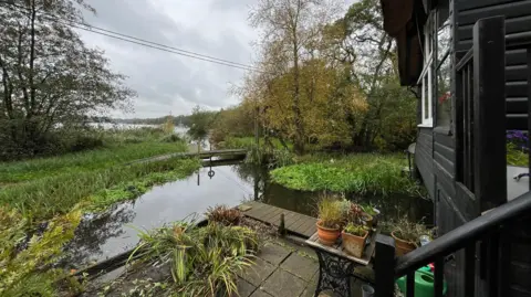 Shaun Whitmore/BBC The view from a Norfolk Broads' black clad wooden building veranda showing the River Bure coming up in front of the house and bearing to its right to go under the house. Trees can be seen further off on either side of the river, which then can be seen joining a larger mass of water