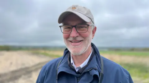 Matthew Norris-Hill standing in front of a field wearing a grey cap, blue rain coat and glasses.
