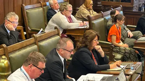 BBC Alfred Cannan is sitting on his seat in the House of Keys chamber, on a green chair. He is wearing a dark suit and tie, he reads from a pile of A4 papers laid out on the desk. There is also a laptop and a glass of water on the desk. Other members of the House of Keys sit nearby and listen on. The seats and benches are wooden and upholstered with avocado green leather. 