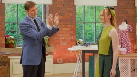 Patrick Grant in a light blue blazer and dark trousers stands with both hands raised, gesturing as if explaining a detail as he faces a model wearing a dark and light green top and trousers in colour blocks. Large windows and sewing materials are visible in the background.