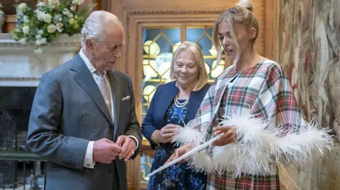 Getty Images The King, is looking at a document held by Siobhan Mackenzie. He is wearing a grey suit and light tie, she is in a white, red and green tartan coat. They are in Dumfries House, an elegant palladium mansion.