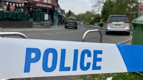 BBC A close-up of police tape sealing off a road with shops and cars