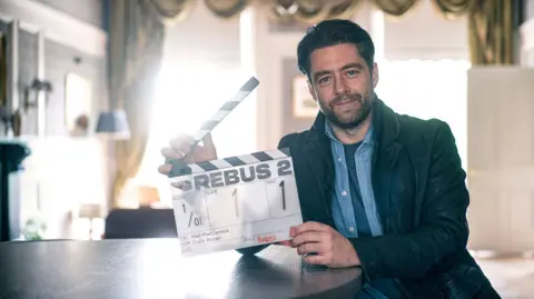 BBC/Eleventh Hour Films/Mark Mainz Richard Rankin. He has dark hair, a beard and a moustache and is wearing a blue denim shirt with a black jacket over the top. He is holding a clapperboard with REBUS 2 in large black letters.