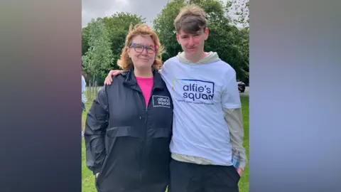 BBC/Mairead Smyth A woman with dark blonde shoulder length hair is wearing a dark coloured jacket with a white logo saying Alfie's Squad. She is smiling while standing next to a teenage boy also smiling and wearing a white Alfie's Squad T-shirt over a pale coloured hooded top. They have their arms around each other. There are trees behind them.