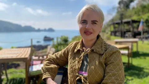 Natalia Silvester has short blonde hair and is wearing a green jacket, sitting on a wooden chair at a cafe overlooking the sea.