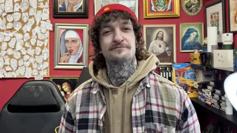 Ross Walker, he is wearing a red hat, light brown jumper and check shirt standing in his tattoo parlour. 