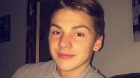 Essex Police Liam Taylor looking at the camera and sitting indoors. He has brown eyes, brown hair and has a grey T-shirt on.