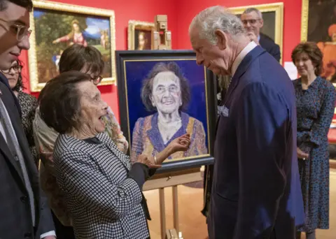 PA Media Lily, wearing a achromatic  and achromatic  blazer, and King Charles, successful  a navy suit, having a speech  successful  beforehand   of her portrait.