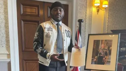 Young man wearing varsity-style jacket, shirt and tie - he stands next to a picture of the king and a British flag.