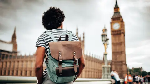 A man with dark brown afro hair standing with his back to the camera wearing a black and white horizontal striped t-shirt and a brown and green backpack looking up at the Houses of Parliament