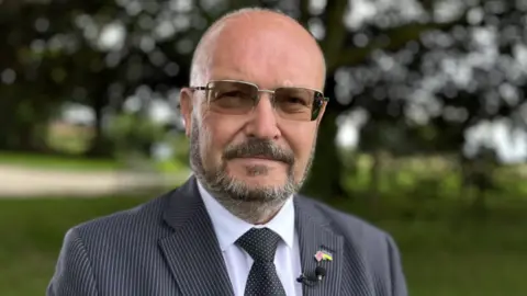 Martin Giles/BBC Graham Plant in tinted-lens spectacles, a pin-striped dark jacket, white shirt and black tie with tiny white spots 