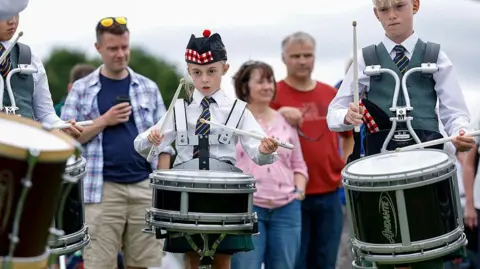 Getty Images Two young drummers in traditional outfits playing drums that are strapped to them. There are spectators behind them