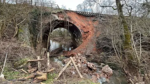 BBC A bridge underneath a railway with a river running underneath it. Earth has fallen away from the railway track leaving it exposed. There is debris and fencing in the river below.