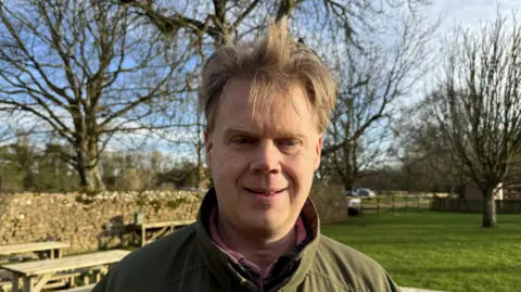 A man with blonde hair wearing a green jacket and pink shirt stood on green grass with a blue sky. 