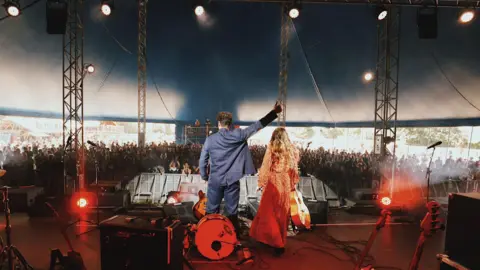 Family photo Ferris and Sylvester wave to the crowd in Glastonbury's Avalon stage after their performance at the 2022 festival, the crowd in the large blue tent is big and numerous instruments are on the stage. The pair have their backs to the camera. 