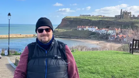 Steve Jones/BBC Neil Orford stood on the cliff top. He is wearing sunglasses and a black hat, with a black gilet and purple jumper.