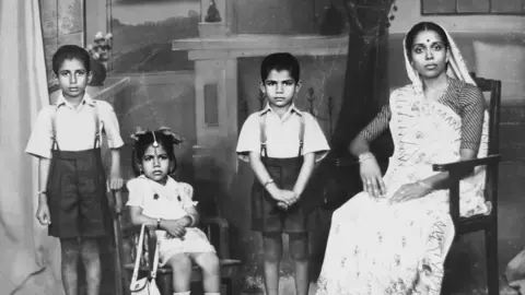 A black and white family portrait with four of Tara Mistry's family members. On the left is a young boy who is standing up wearing a short-sleeved shirt and black shorts. To his left, a young Tara Mistry is sitting down on a chair wearing a white dress. In the middle is another boy, standing and also wearing a shirt and shorts. On the far right is an older woman who is sitting down wearing a saree.