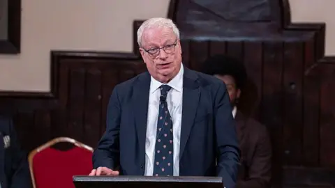 Getty Images Lord Chris Smith wearing a navy blue suit and white shirt with matching navy blue tie with the Cambridge University crest. He has white hair and black rimmed glasses. He is standing behind a podium.