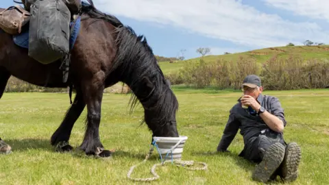 sesmlphotography Roger Sewill sitting on the grass during his hill forts trek, drinking beside his Dales pony Scarlet. She is also drinking from a bucket in front of her. It is a sunny day.