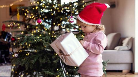Child opens Christmas presents in a santa hat infront of a Christmas tree