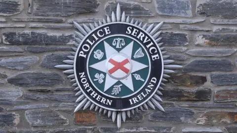 A PSNI crest on a grey stone wall. The crest reads "Police Service of Northern Ireland" and has a red cross in the middle surrounded by various symbols, including a harp, shamrock, crown, torch and scales (of justice). 