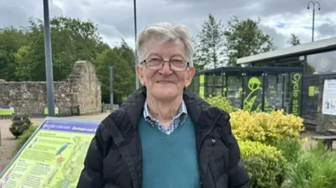 A man with grey hair, wearing a black jacket, blue v neck jumper and shirt is standing facing the camera and smiling. Behind him are some bushes and a cycle shop.