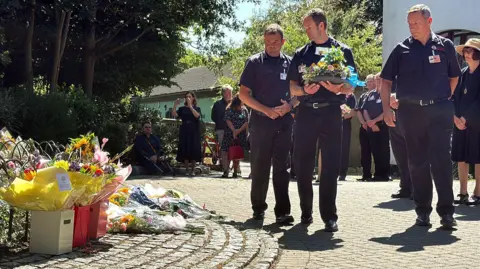 Essex Police Three male firefighters, one carrying a bouquet of flowers approach an area in the park where flowers and soft toys have been left in a circle in tribute to the deceased child Leonna Ruka who died after a falling tree branch collapsed on her. Several men and women can be seen standing nearby. It is a warm and sunny day.