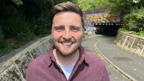 Tom Deakin is smiling at the camera. He is standing on a path leading to the entrance to the underpass. The bridge above it has been sprayed with graffiti.