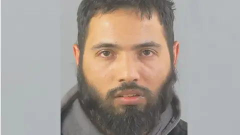 A custody photo of Sohail Amiri who has dark hair and a full beard and wears a hooded top