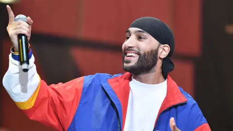 BBC Manni Sandhu, a male artist wearing a blue and red jacket with a white top, holding a black microphone while looking to his right