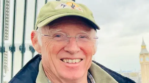 Family handout/TVP Peter Hughes is wearing a green cap and a blue coat and green jumper, with the Elizabeth Tower in London on the right of the picture