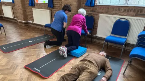 A lady wearing a pink jumper kneeling on a cushion alongside a man wearing brown trousers and a brown jumper who is lying on the floor
