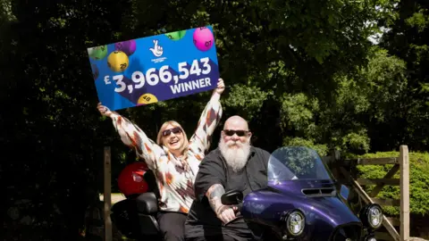 A woman holding a sign that says "The National Lottery and £3,966,543 winner". The background of the sign in clue and there multicoloured bingo balls. There is a man sat in front of the woman on a purple Harley-Davidson trike bike. There are trees behind them and a wooden fence.  