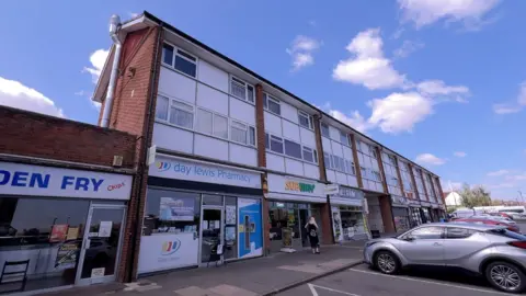 Day Lewis Pharmacy, situated in the middle of a parade of shops with Subway on one side and a fish and chip shop on the other. There are two floors of residential flats on top of the pharmacy with white cladding around the windows. There are several cars parked in front of the parade of shops