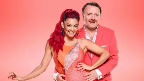 BBC/PA Comedian Chris McCausland, wearing an open-necked white shirt and a pink jacket, holds the waist of dancer Diane Buswell, who has long dyed bright red hair and who is wearing a brightly-coloured sparkly leotard, in a promotional shot.